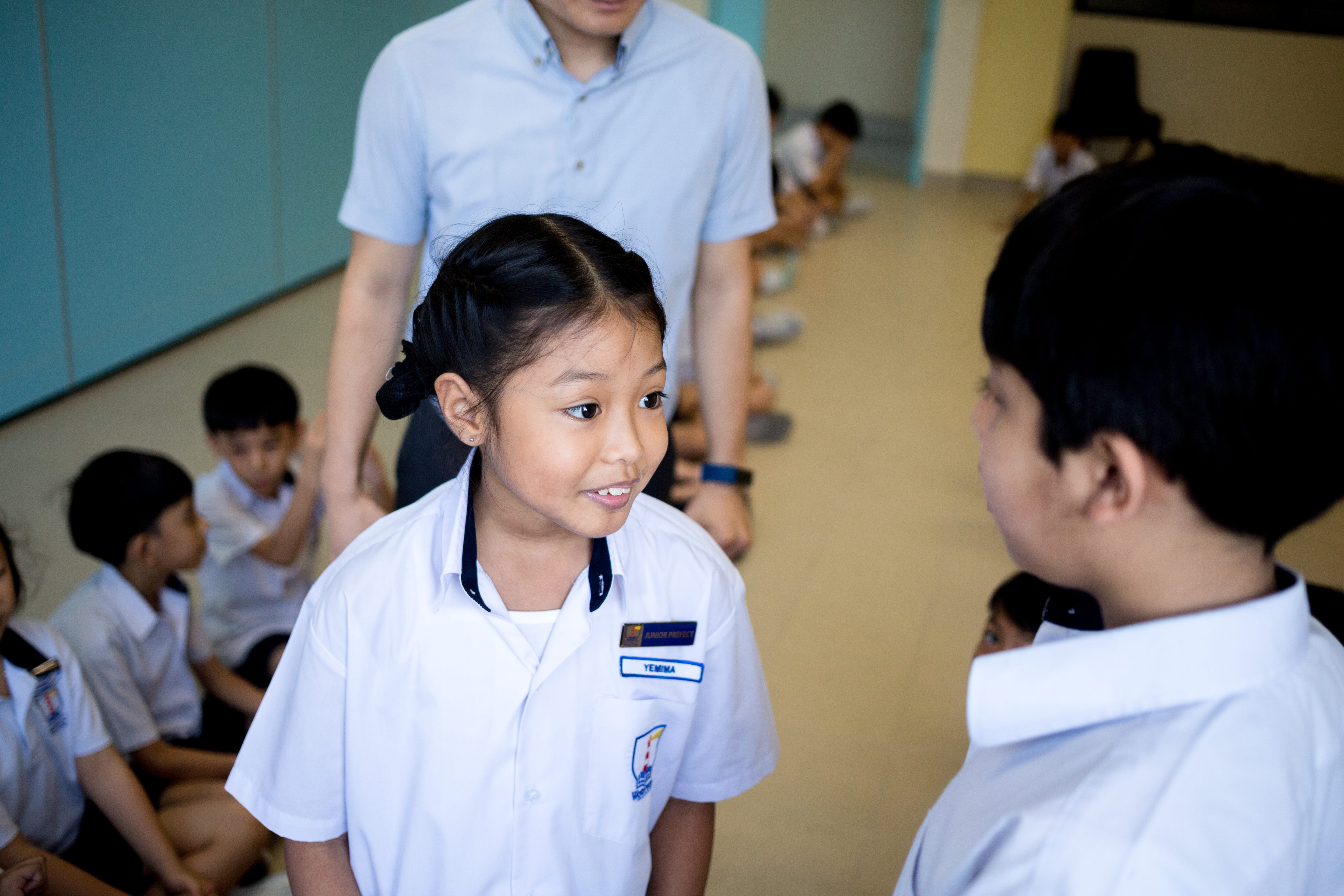 Schoolgirl in white uniform talking to a boy, with other students seated on the floor behind.
